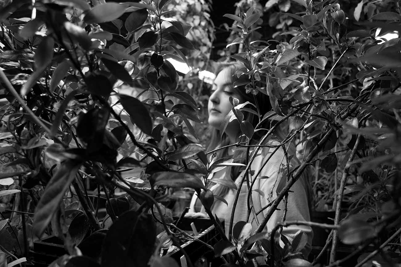 Black and white, Girl standing peacfully obscured by rows of vines and plants in greenouse