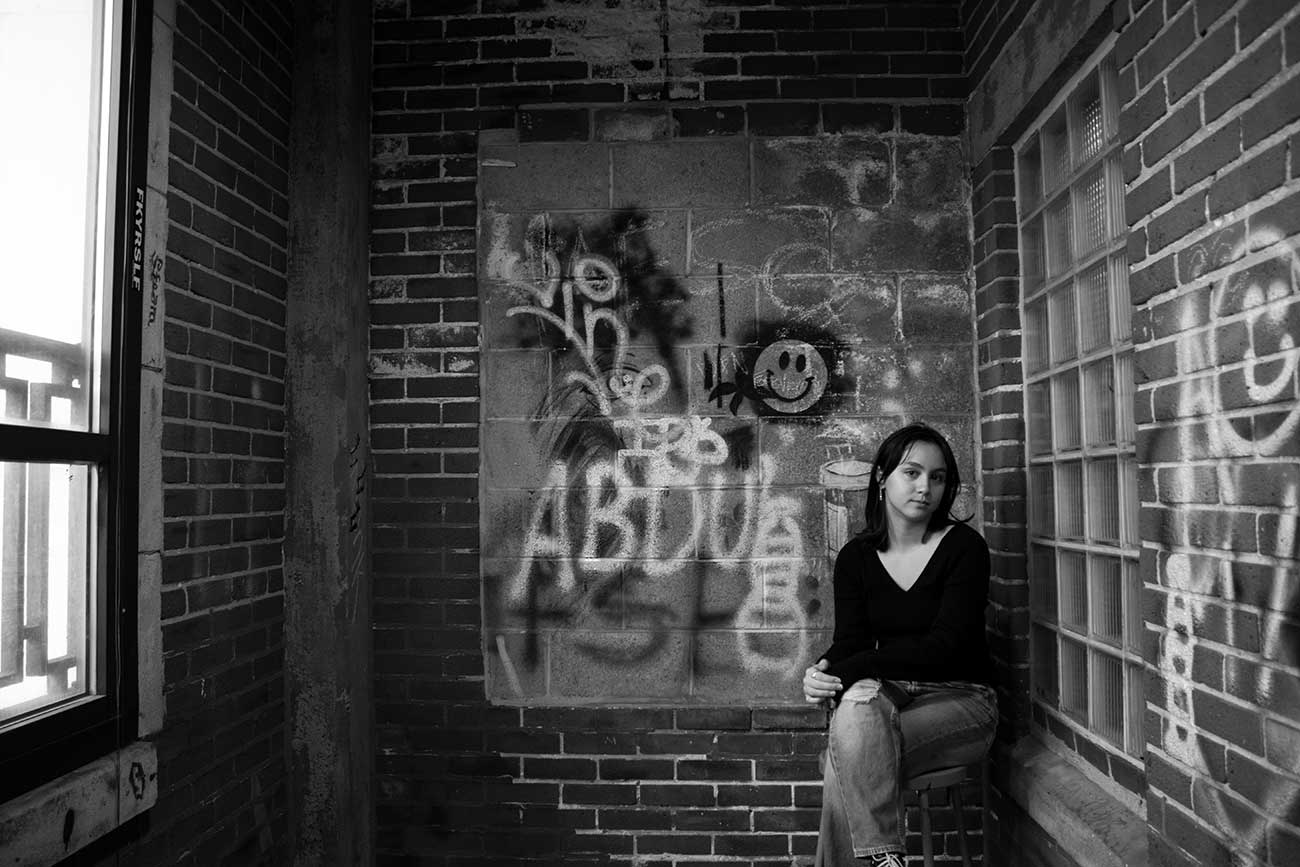 Black and white photo of a girl sitting on a still in a graffitied brick room